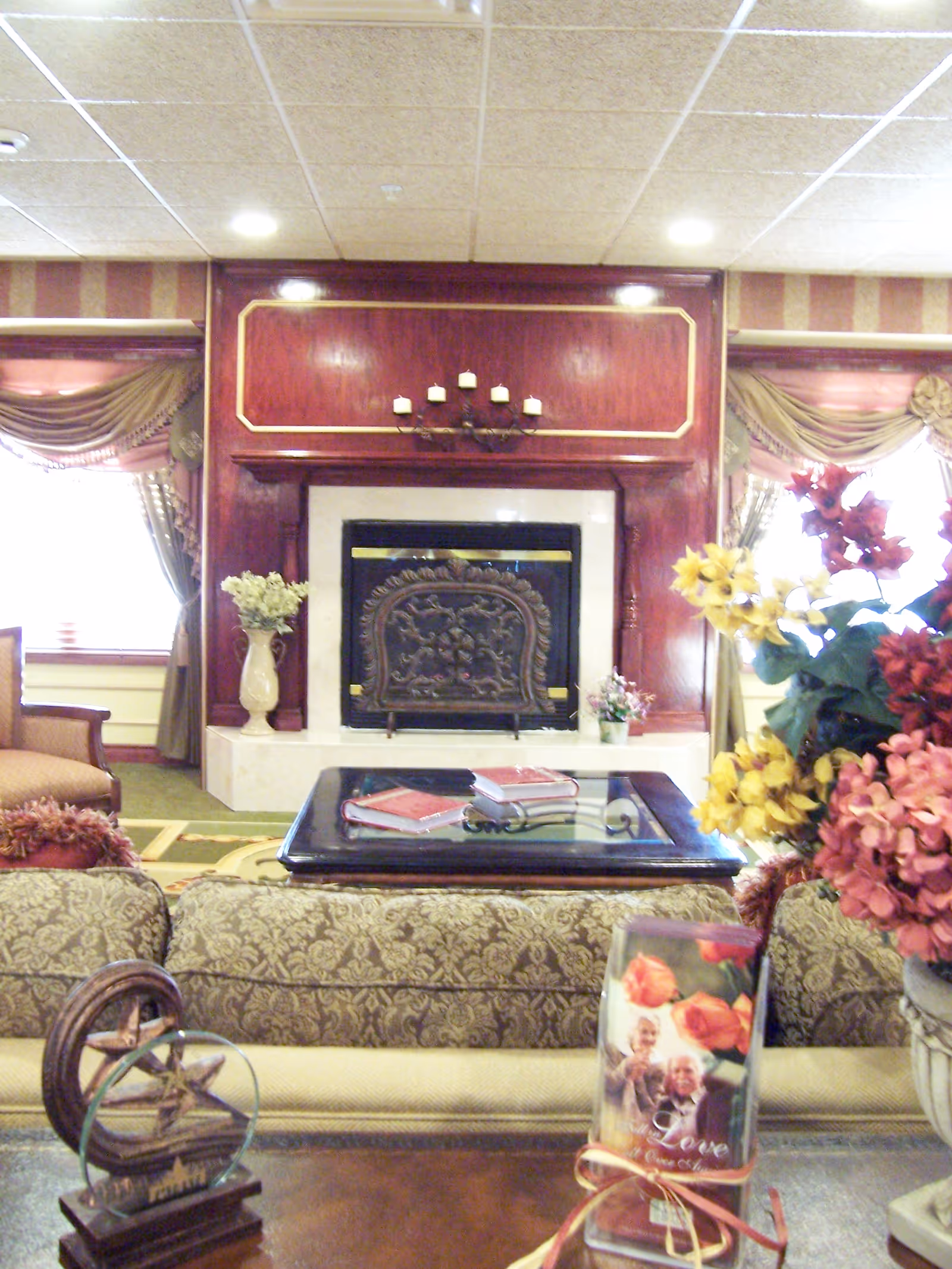 A cozy living room area with a decorative fireplace in the center, flanked by windows with elegant drapes. In front of the fireplace is a glass-top coffee table with books on it. The foreground shows a patterned sofa and a table with a floral arrangement and a decorative item featuring an elderly couple and the word 'Love'.