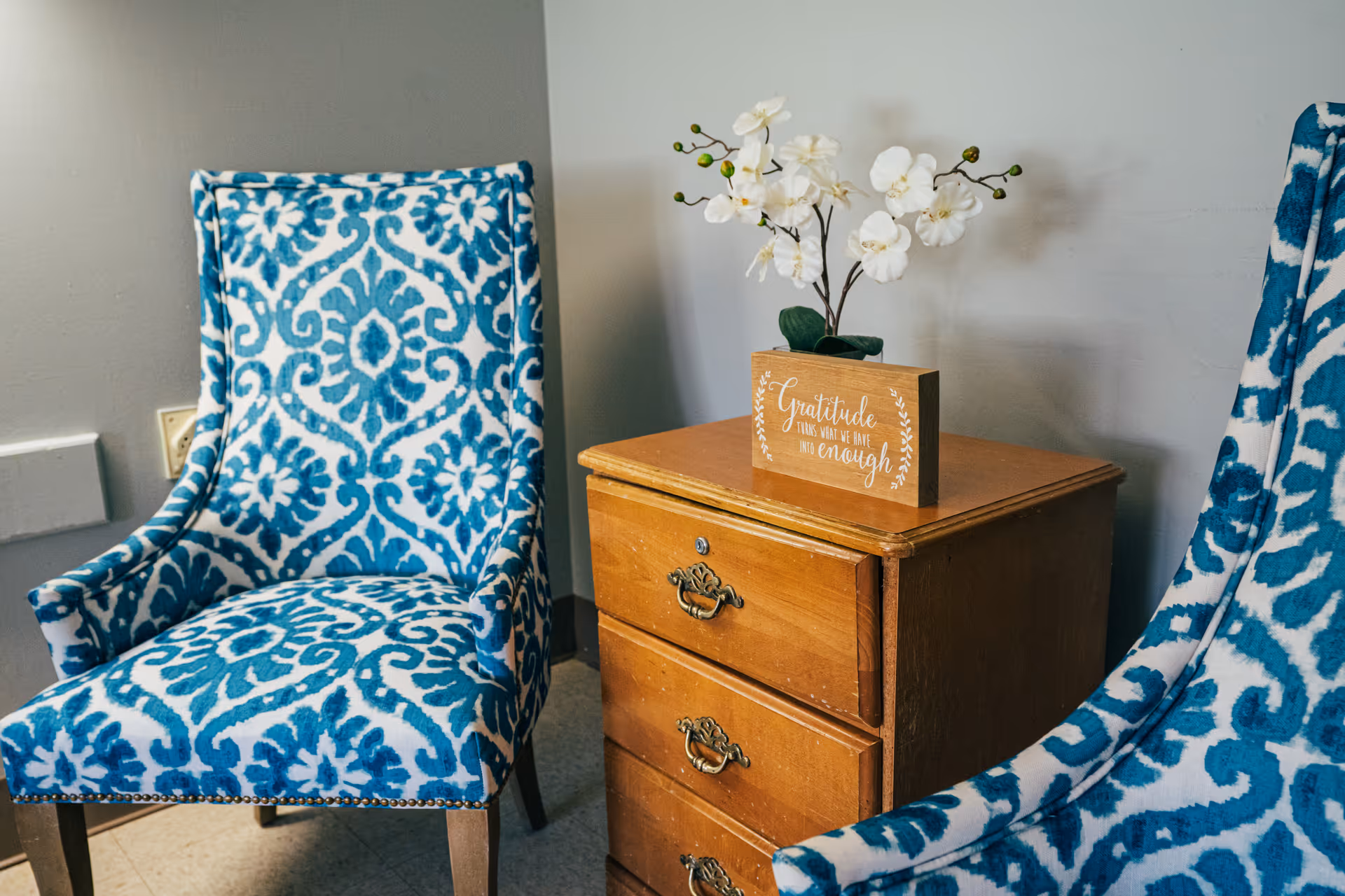 Two blue and white patterned upholstered chairs positioned on either side of a wooden three-drawer nightstand. On top of the nightstand is a small wooden sign with the word 'Gratitude' and a white orchid plant. The setting appears to be a cozy indoor room with a light gray wall in the background.
