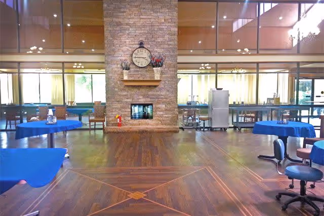 Spacious interior room with wooden flooring and a large stone fireplace in the center. The fireplace has a clock and decorative plants on the mantel. The room has several tables covered with blue tablecloths and chairs arranged around them. Large windows with curtains allow natural light to enter the room.