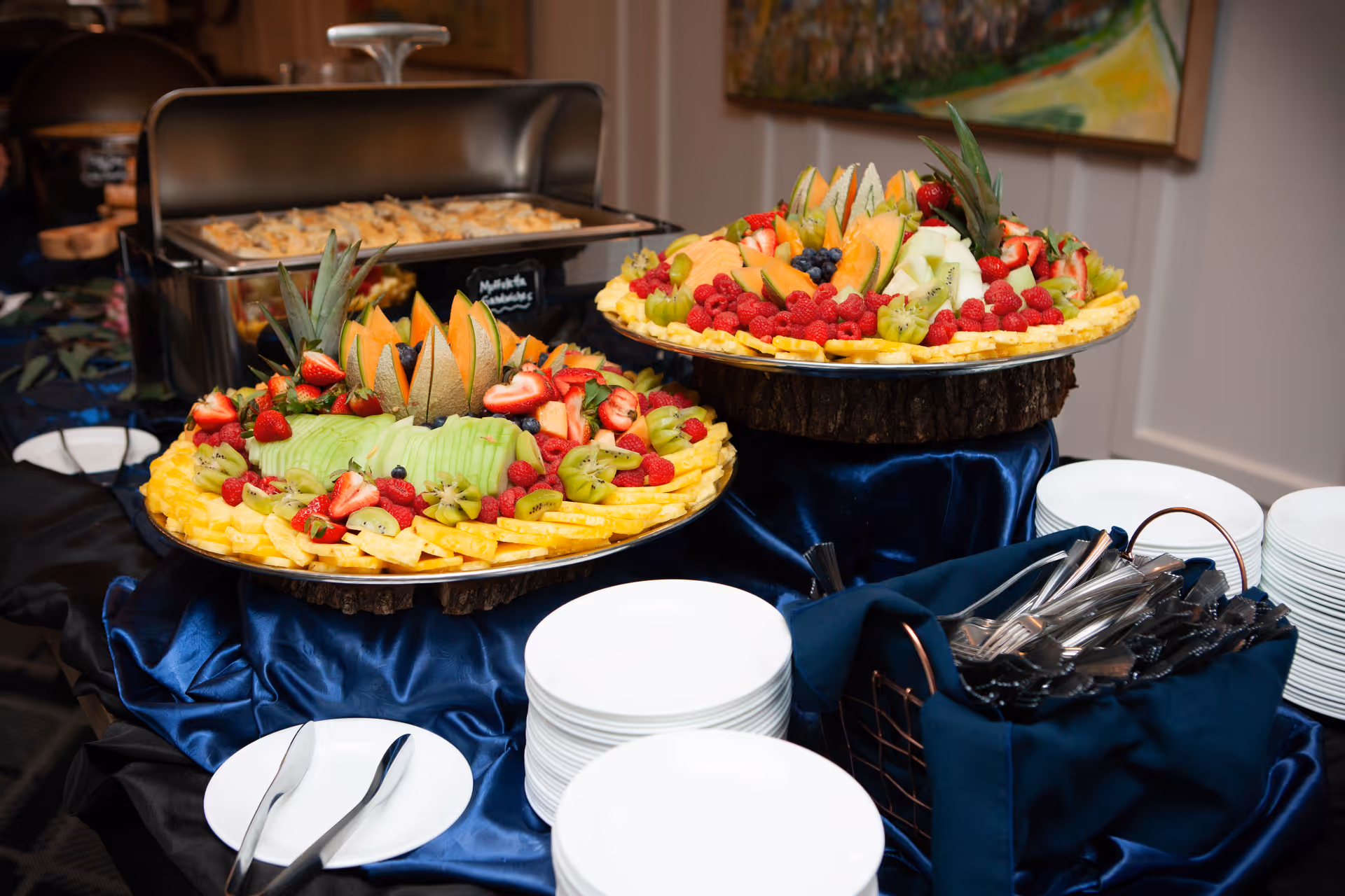 A buffet table displaying two large decorative fruit platters with stacks of plates and utensils.