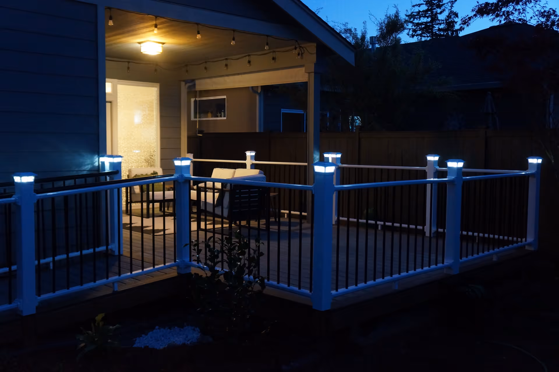 A lit outdoor deck at dusk with white railings and illuminated post caps and patio furniture under a covered porch.
