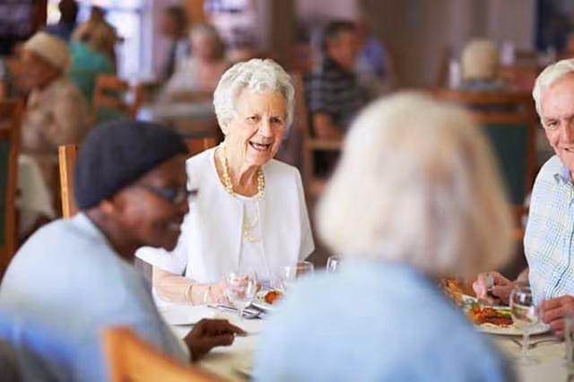 A group of elderly people sitting around a dining table, enjoying a meal and engaging in conversation in a bright dining room setting.