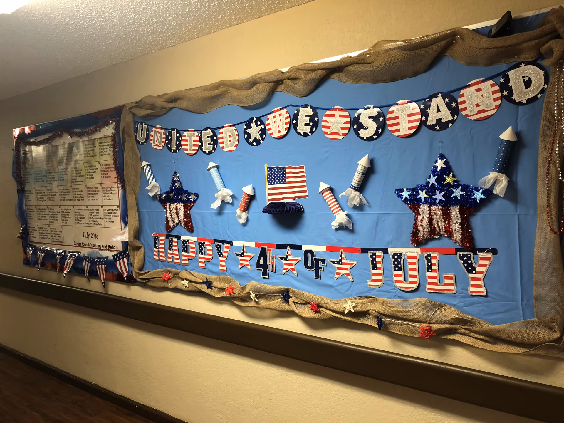 Hallway bulletin board decorated for the Fourth of July with American flags, stars, rockets, and 'Happy 4th of July' lettering.