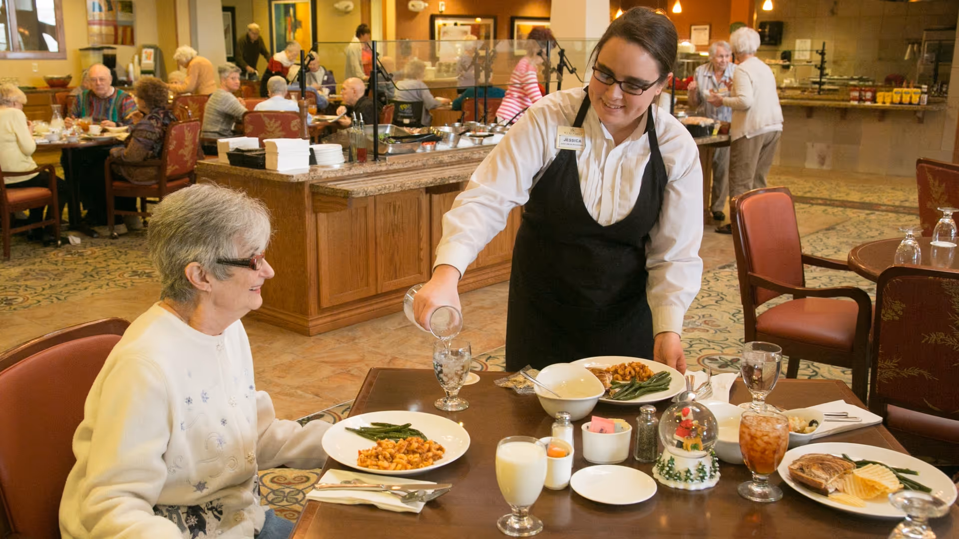 A server pours water for an elderly woman seated at a table in a retirement community dining room while other residents eat in the background.