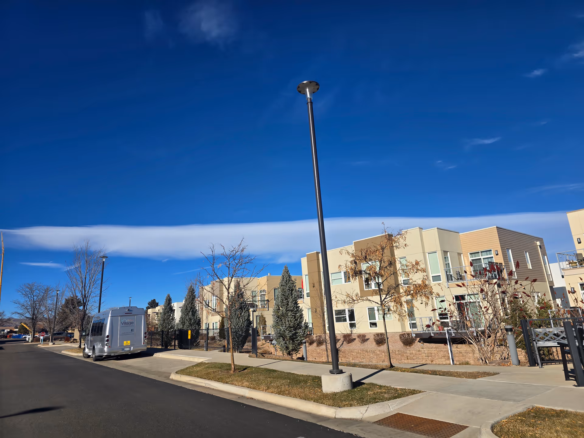 Street-level view of a modern multi-story senior living building with a parked shuttle van, lampposts, trees, and a clear blue sky.