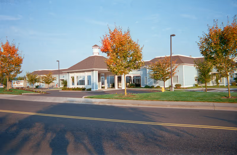 Exterior view of a single-story building with light blue siding and a brown roof, surrounded by trees with autumn foliage. The building has a covered entrance and a sign that reads 'Cascade Care Center'. The scene is set on a clear day with a blue sky.