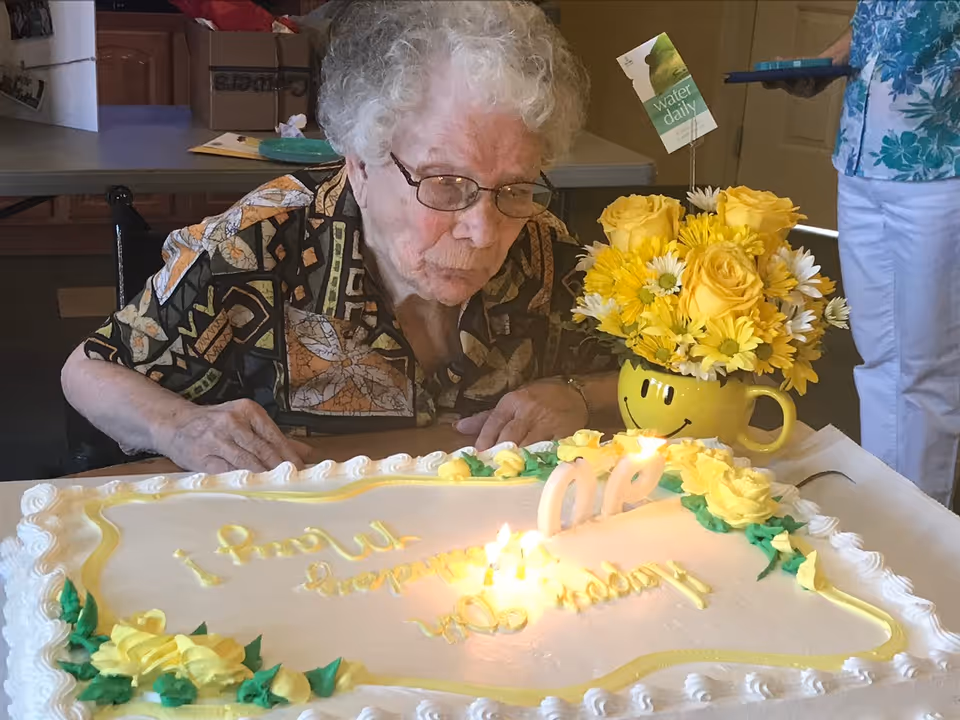 An elderly woman with glasses and curly white hair is sitting at a table, leaning forward to blow out candles on a large birthday cake decorated with yellow flowers. Next to the cake is a yellow smiley face mug filled with a bouquet of yellow and white flowers. A person wearing white pants and a blue floral shirt stands nearby.