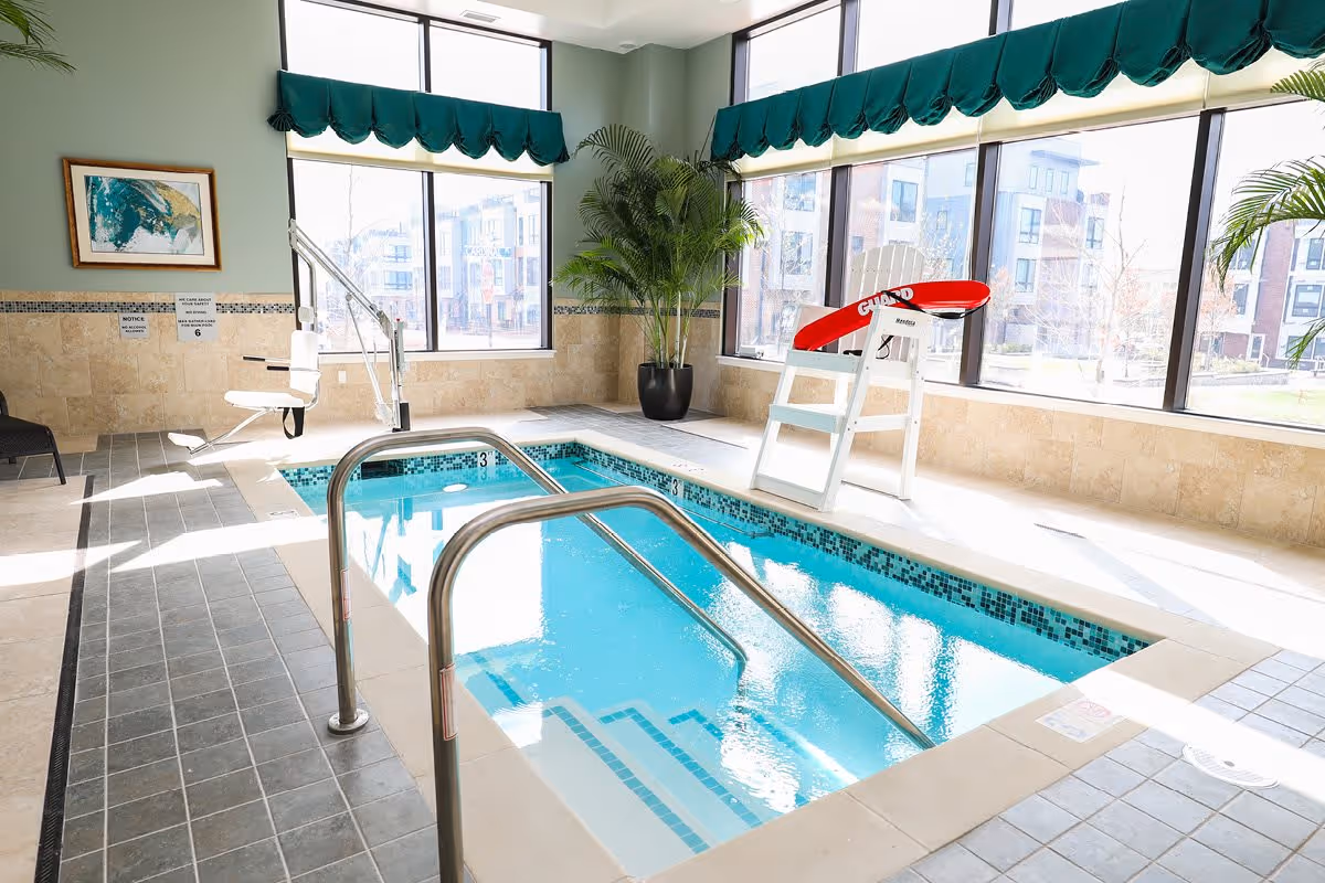 Indoor swimming pool area with large windows letting in natural light. The pool has metal handrails and a lifeguard chair with a red seat. There are green plants in pots and a framed artwork on the wall. The walls are tiled halfway up with beige tiles and the floor has gray tiles.