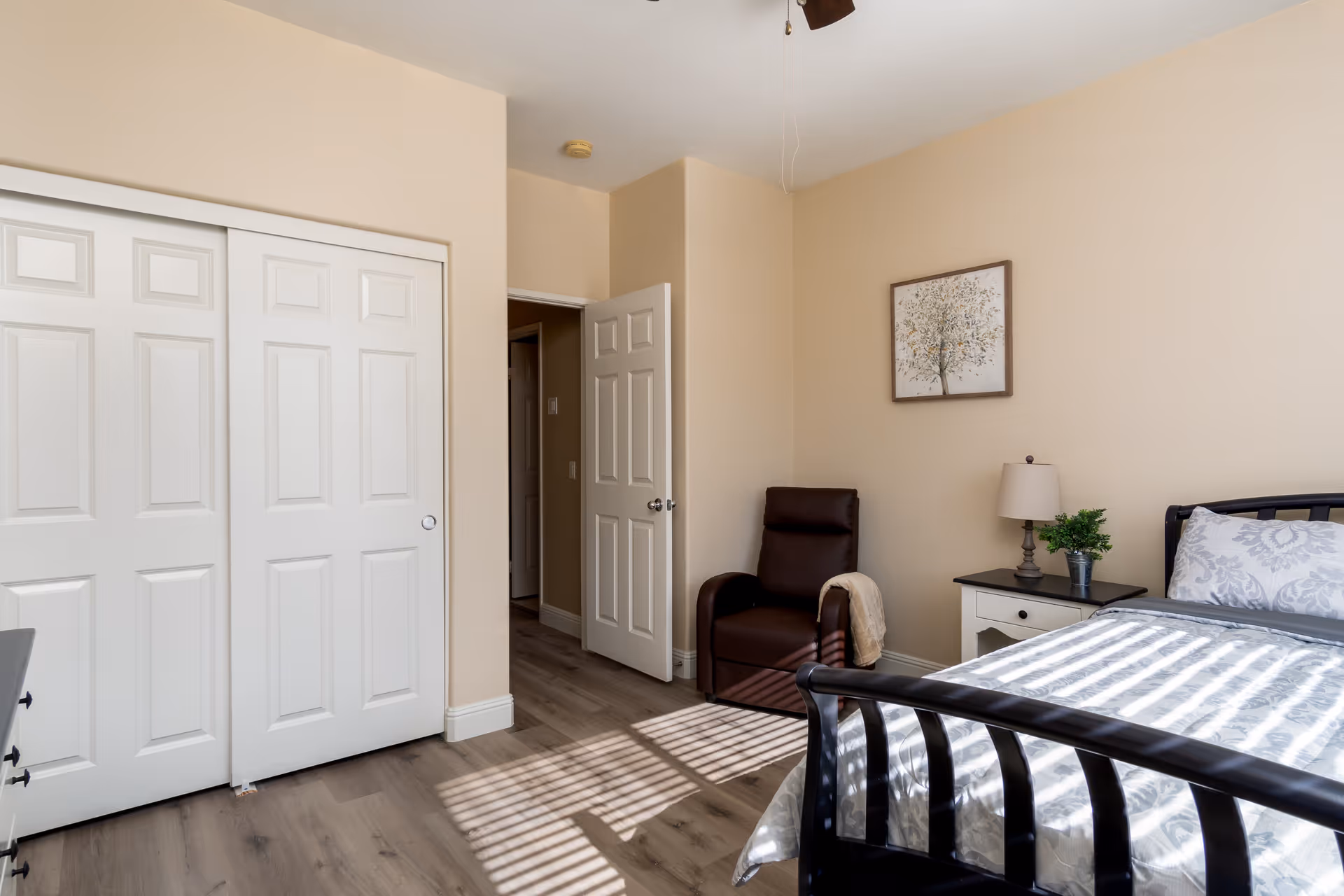A bright bedroom with beige walls and wood flooring. The room features a black metal bed frame with a patterned white and gray bedspread, a white nightstand with a lamp and a small potted plant, a brown leather armchair with a beige throw draped over one arm, and a framed picture of a tree on the wall. There is a closet with white sliding doors and an open door leading to another room.