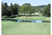 A scenic view of a golf course with a well-maintained green, sand bunkers, a small water hazard, and trees in the background under a clear sky.
