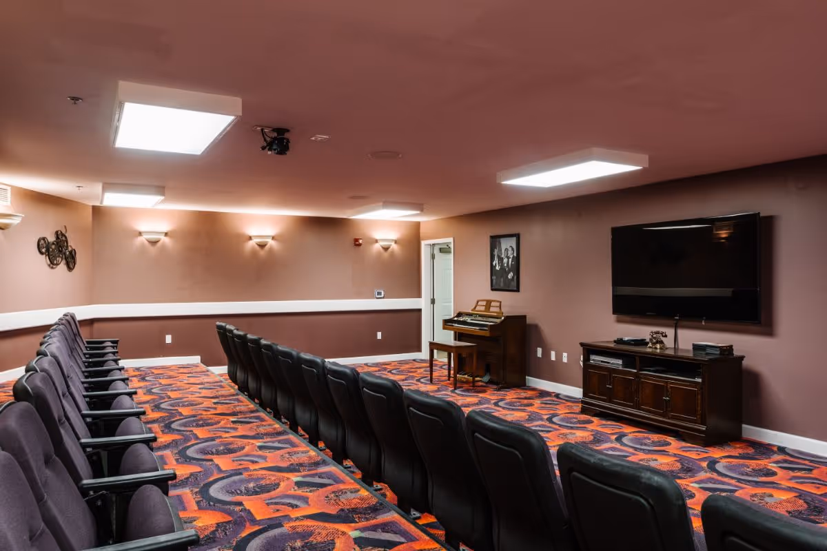 A small theater or media room with two rows of black cushioned chairs facing a large flat-screen TV mounted on a wall. The room has patterned carpet in shades of orange, purple, and black, mauve-colored walls, and ceiling lights. There is a wooden piano with a bench against the far wall and a black and white framed photo above it.