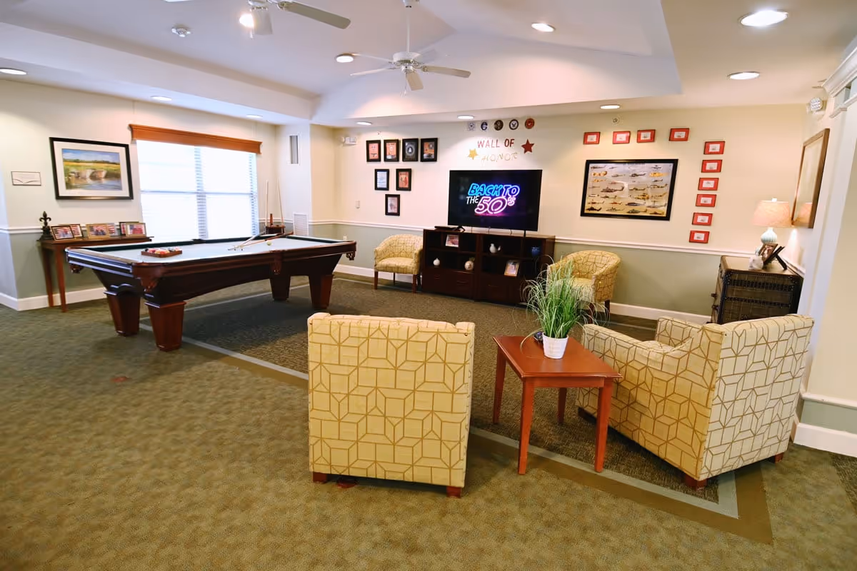 A senior living common room featuring a pool table, a TV on a media console, and a seating area of patterned armchairs around a small table.
