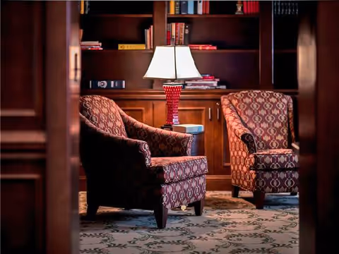 Two patterned armchairs with a small table and a lamp between them, set in front of a wooden bookshelf filled with books in a cozy interior space.