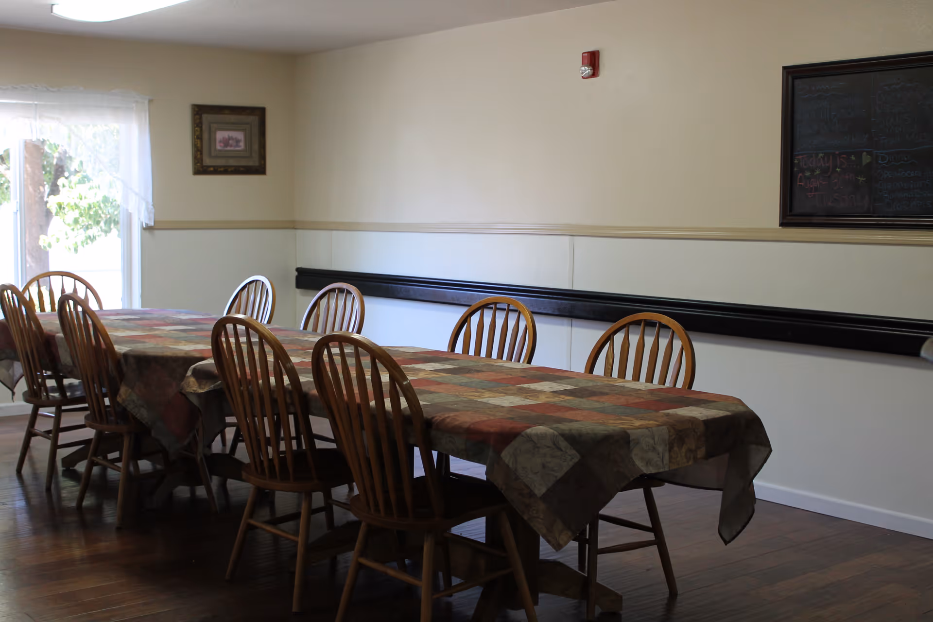 A dining room with a long rectangular table covered with a patchwork-style tablecloth. There are eight wooden chairs around the table. The room has light-colored walls, a window with sheer white curtains on the left, a framed picture on the wall, and a chalkboard on the right wall.