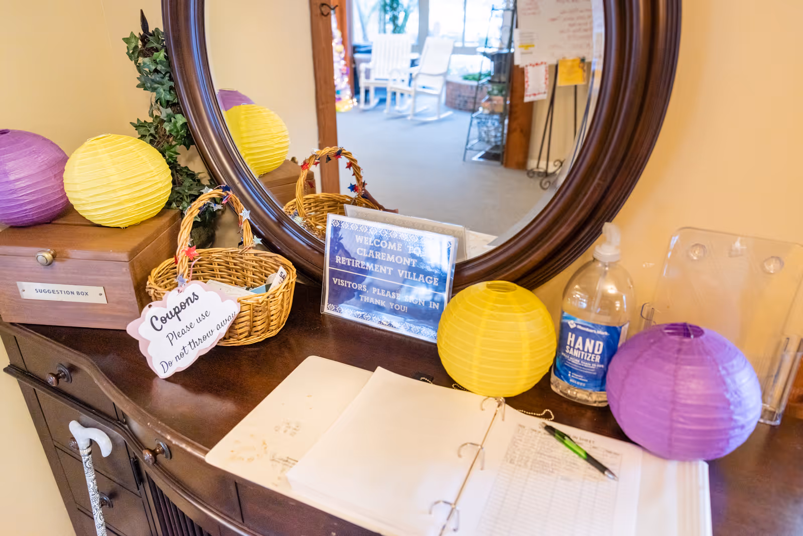 A wooden console table with a large round mirror above it in a retirement village. On the table are two small wicker baskets, one with a sign that says 'Coupons Please use Do not throw away', a bottle of hand sanitizer, two paper lanterns in yellow and purple, a suggestion box, and an open binder with a pen. In the mirror's reflection, a hallway with chairs and a bulletin board is visible.