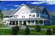Large light-gray two-story senior living building with a wraparound porch, turret, flag, and landscaped lawn under a blue sky.