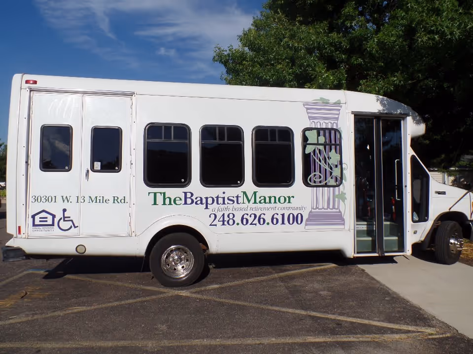 White shuttle bus parked in a lot with signage for The Baptist Manor, a faith-based retirement community, including their phone number and address. The bus has a wheelchair accessibility symbol and a graphic of a classical column with ivy.