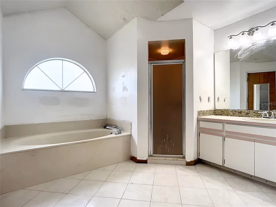 A bathroom with a beige bathtub under a frosted arched window, a separate shower with a frosted glass door, a large mirror above a white vanity with granite countertop, and tiled floor.