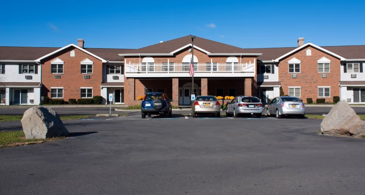 Front exterior view of a two-story senior living community building with a brick facade and white trim. There are four cars parked in front of the building, an American flag on a pole in the center, and large rocks on either side of the parking area. The sky is clear and blue.