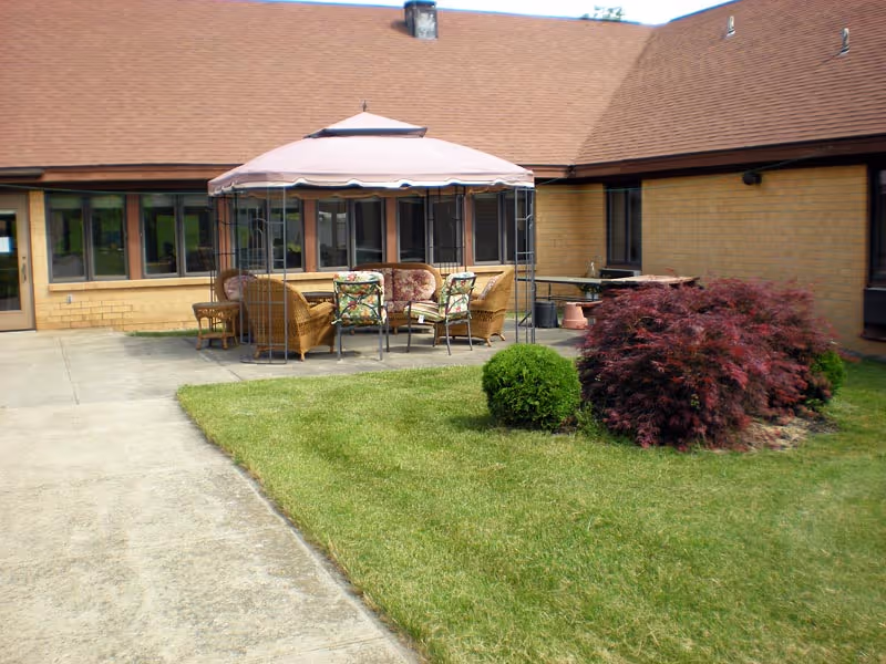 Outdoor courtyard with a canopy-covered patio seating area in front of a single-story brick building, surrounded by lawn and shrubs.