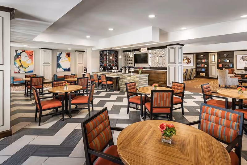 A bright and spacious dining area in a senior living facility with round wooden tables and chairs featuring orange and striped cushions. The floor has a modern geometric pattern in shades of gray and white. In the background, there is a bistro counter with stools, decorative wall art, and bookshelves with various items. The room is well-lit with recessed ceiling lights.