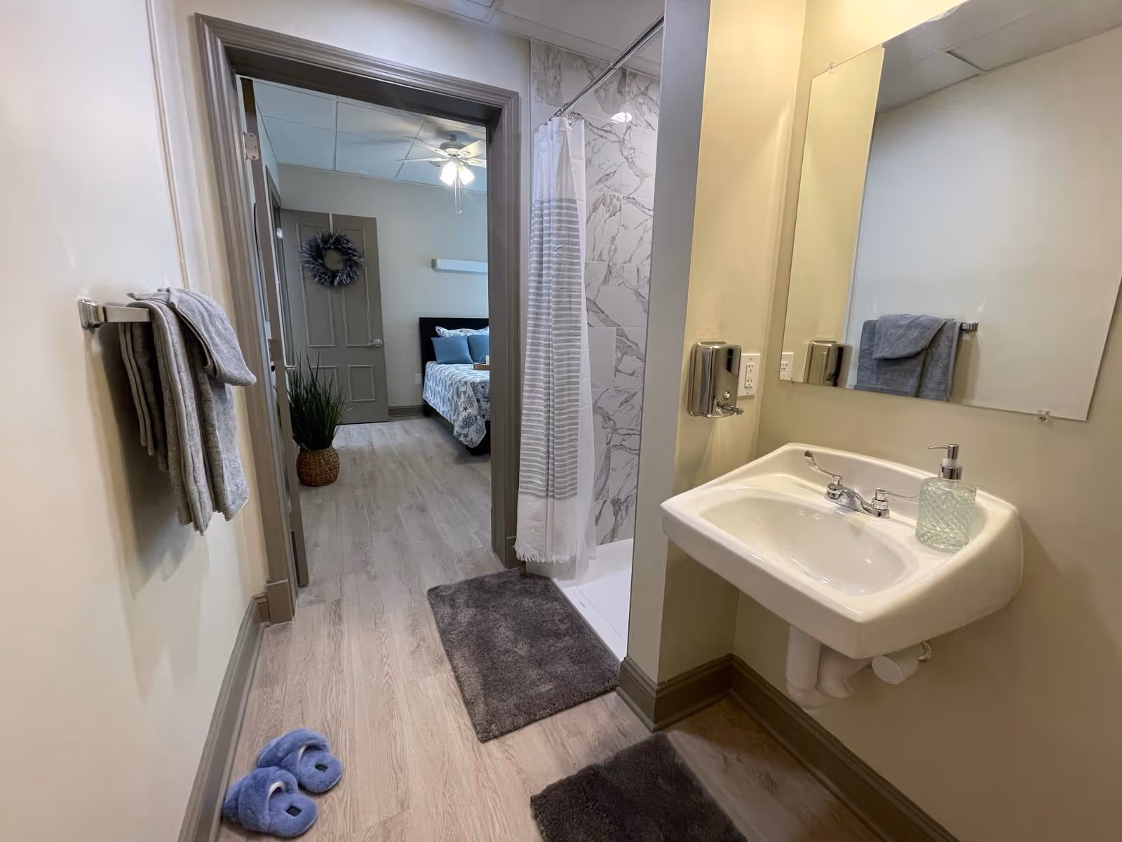 View of a bathroom with a white sink, soap dispenser, and a large mirror. There is a shower with a white and gray striped curtain and marble-patterned wall tiles. Gray towels hang on a rack on the left wall, and blue slippers are on the light wood floor. Through an open door, a bedroom with a bed, pillows, a ceiling fan, and a decorative wreath on the door is visible.
