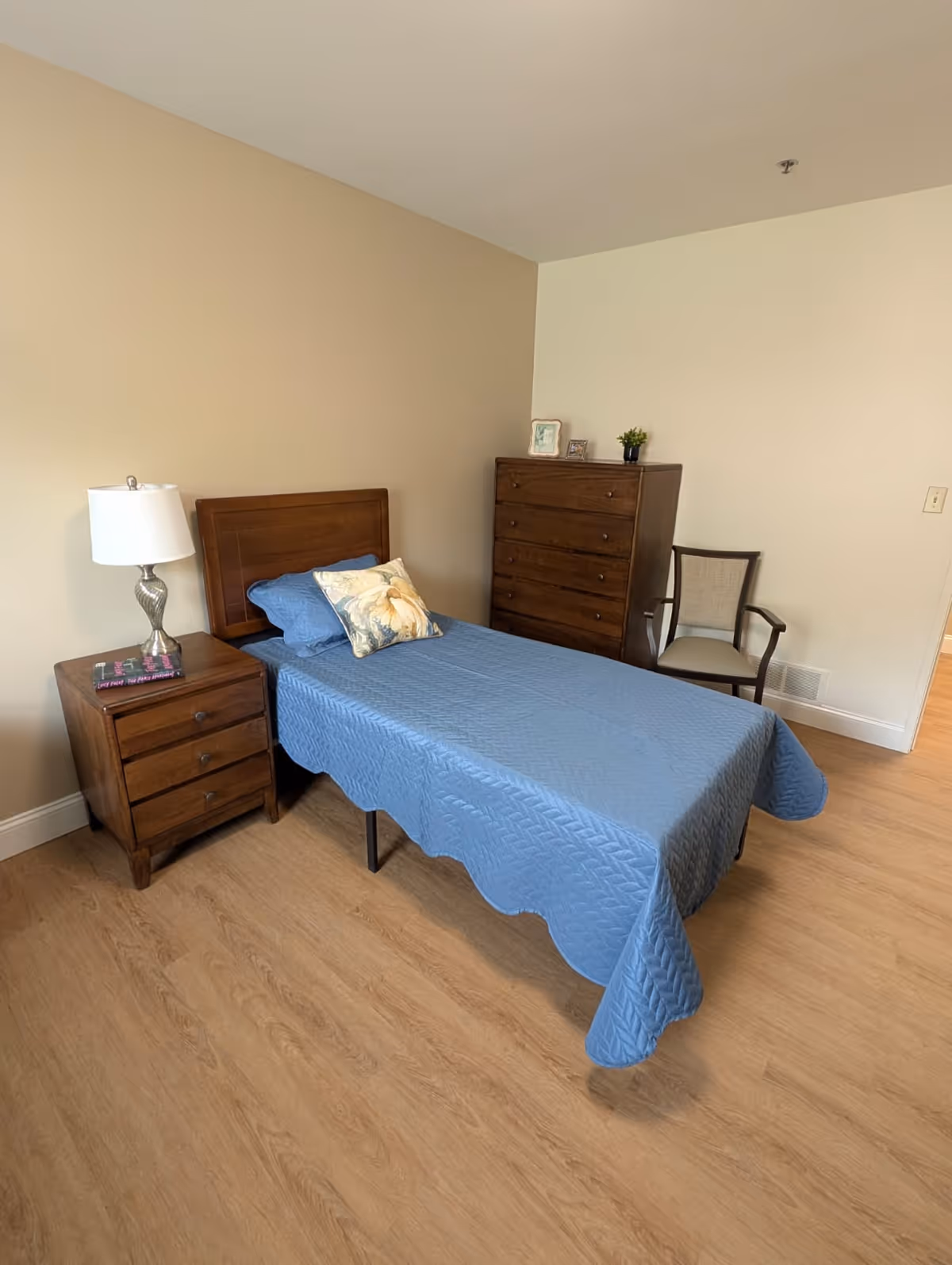 A simple bedroom with a single bed covered in a blue quilt and a floral pillow. Next to the bed is a wooden nightstand with a silver lamp and a book. Against the wall is a tall wooden dresser with small decorative items on top, and a wooden chair with a cushioned seat is placed beside it. The room has light-colored walls and wood flooring.