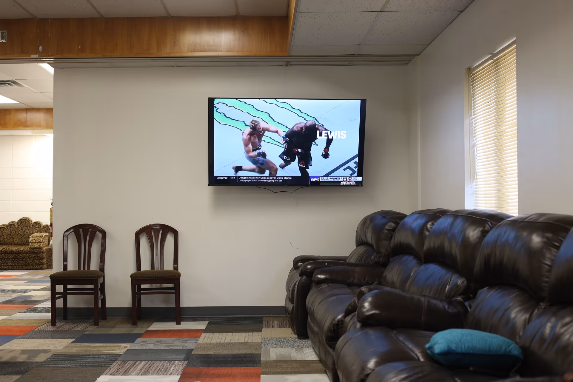 A room with a wall-mounted flat-screen TV showing a mixed martial arts fight. Below the TV are two wooden chairs with cushioned seats. To the right, there is a row of dark brown leather recliner sofas with a blue pillow on one of them. The floor has a multicolored patterned carpet, and there is a window with closed blinds on the right wall.