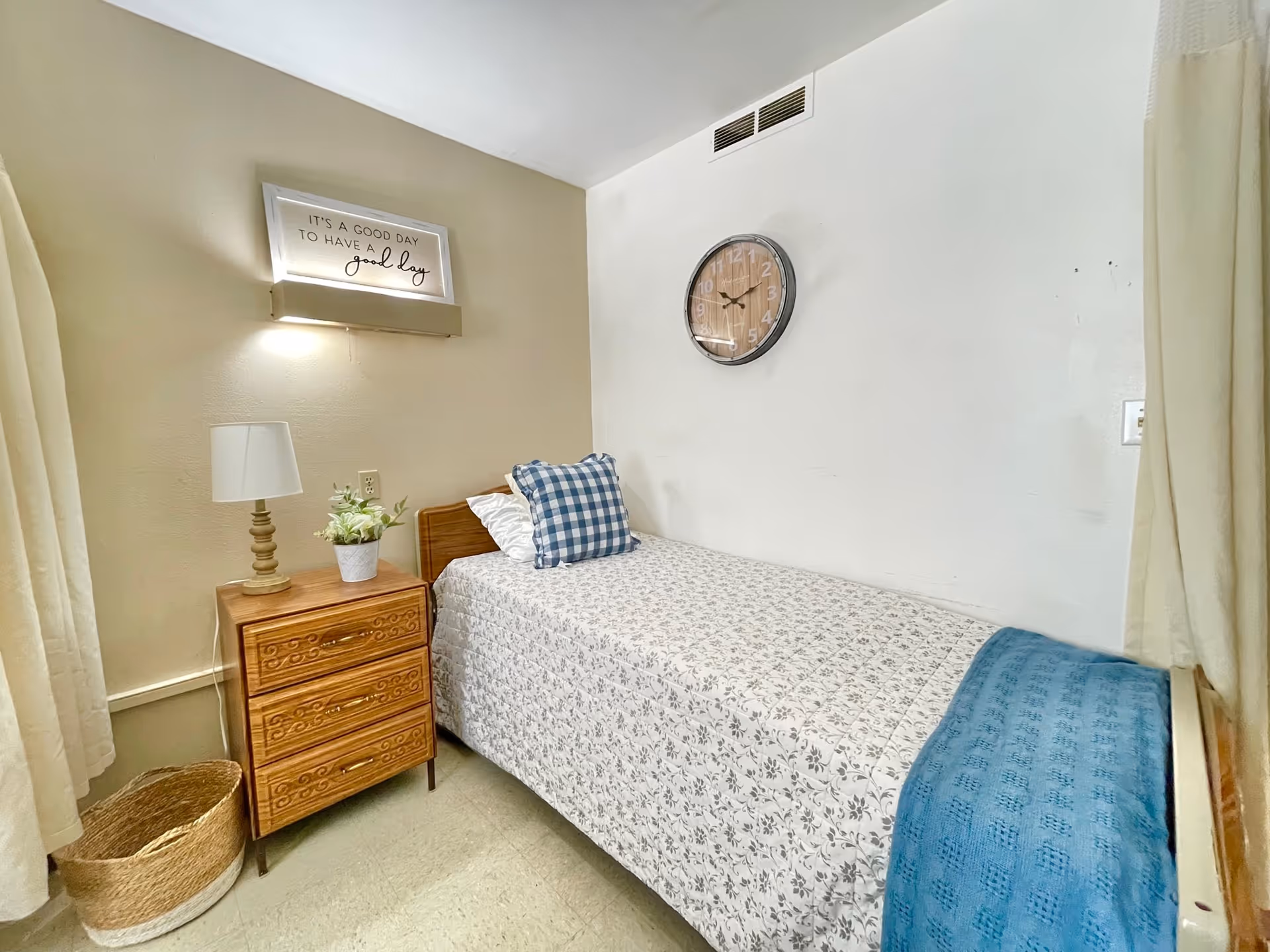 A small, cozy bedroom with a single bed covered in a floral bedspread and a blue throw blanket. There is a wooden nightstand with a lamp and a small potted plant. Above the nightstand, a wall-mounted light illuminates a framed sign that reads 'It's a good day to have a good day.' A round wall clock is mounted on the white wall above the bed. Light-colored curtains frame the window.
