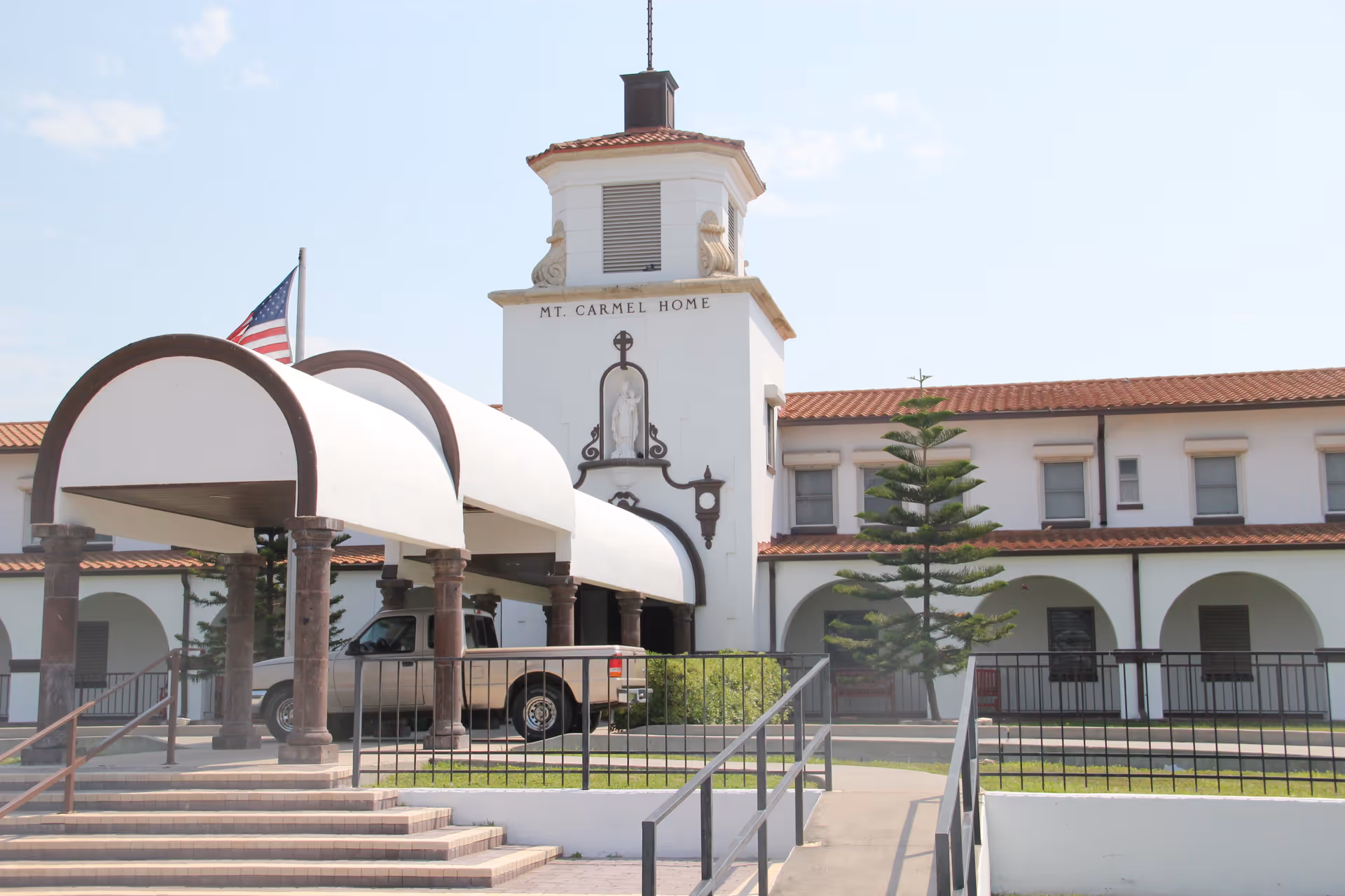 Entrance of the Mount Carmel Home building with a covered drop-off canopy, tower, American flag, and a parked pickup truck.