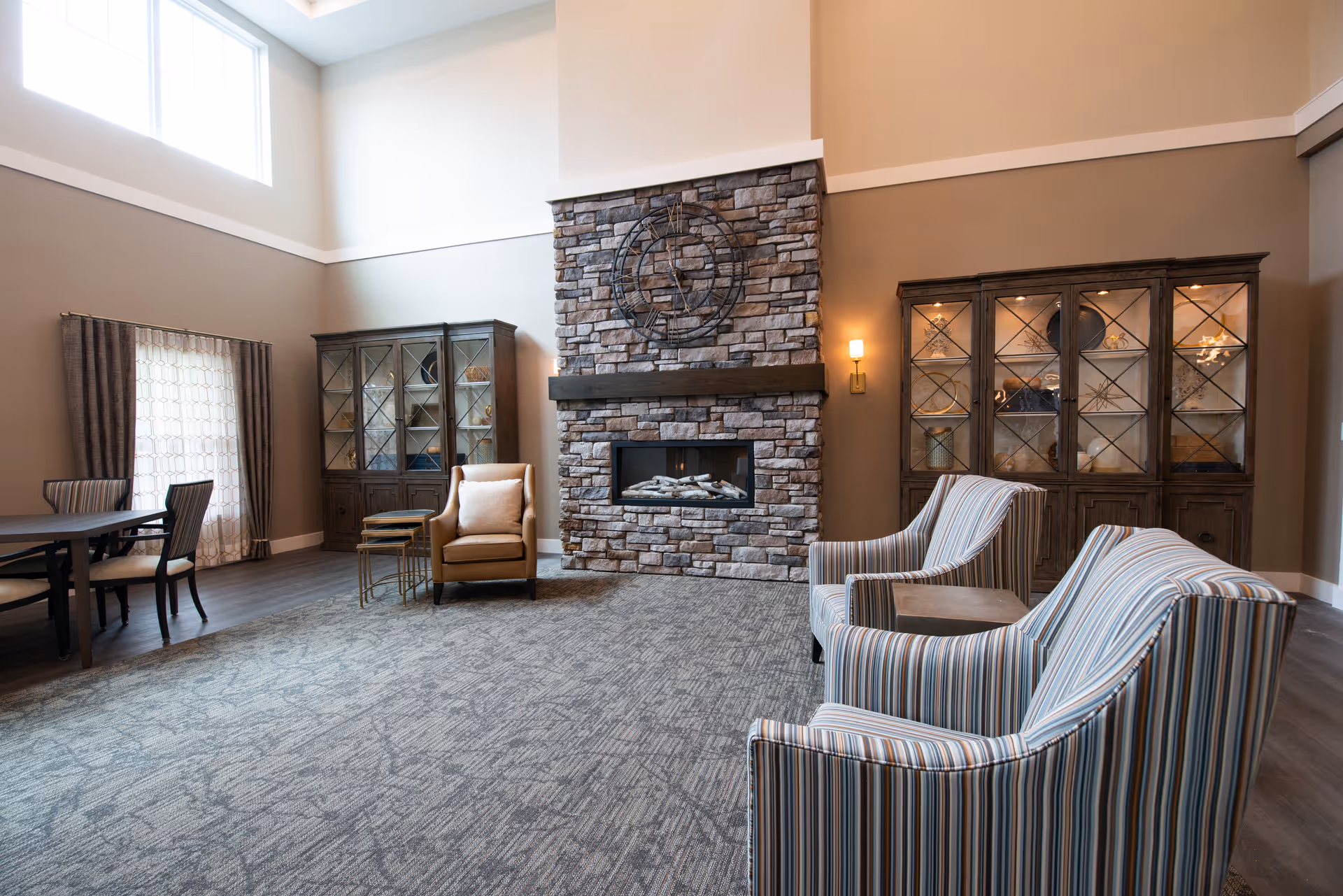 A spacious and well-lit living room area with a high ceiling, featuring a stone fireplace in the center with a decorative metal wall art above it. There are two striped armchairs and a small table in the foreground, a single beige armchair with nested side tables near the fireplace, and two wooden display cabinets with glass doors on either side of the fireplace. A dining table with chairs is visible on the left side near a window with curtains.
