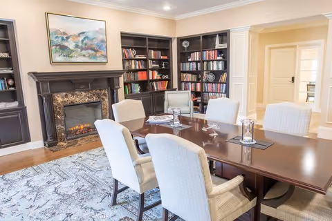 A cozy meeting or dining room with a dark wooden table surrounded by six upholstered chairs. The room features a fireplace with a marble surround and a colorful abstract painting above it. Built-in dark wood bookshelves filled with books and decorative items line the walls. The floor has a patterned area rug, and there is a doorway leading to another room in the background.