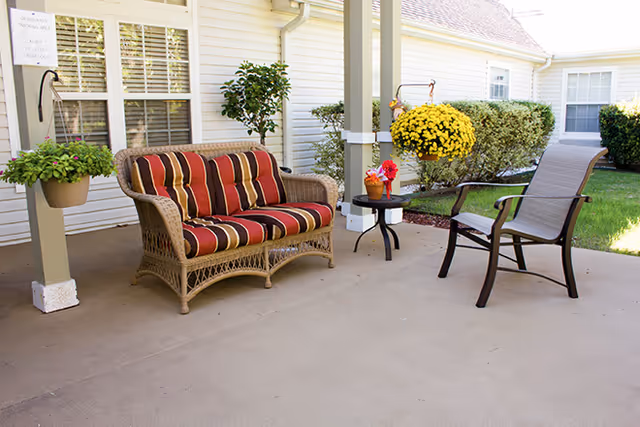 Covered patio with a wicker loveseat with striped cushions, a chair, a small side table and potted flowers beside a building.