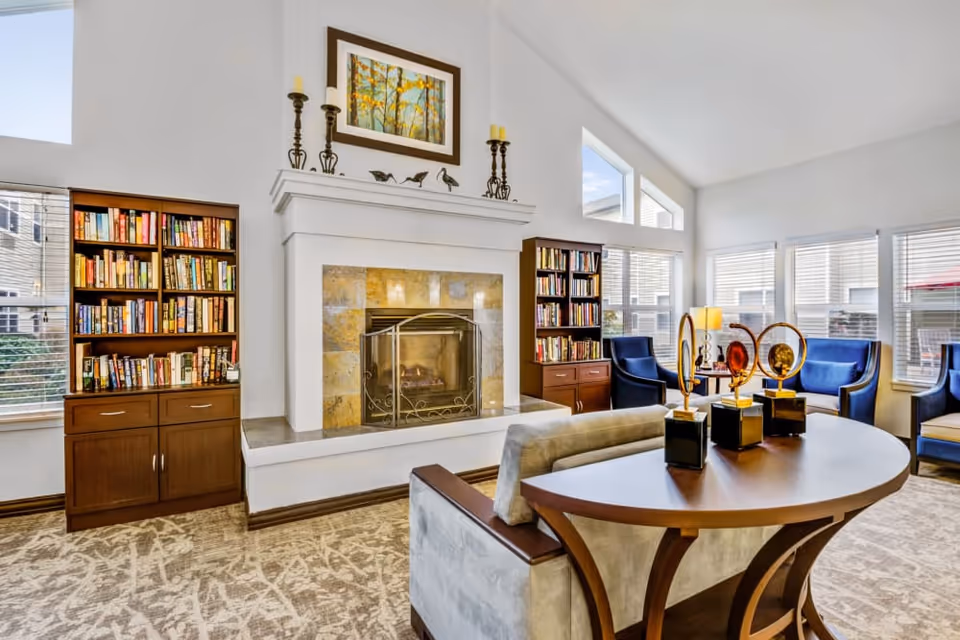 A bright and spacious living room with a fireplace in the center, flanked by two wooden bookshelves filled with books. The room has large windows allowing natural light to fill the space. There are blue upholstered chairs arranged around a small table with a lamp, and a curved wooden table with decorative art pieces in the foreground. The walls are white, and the ceiling is vaulted.