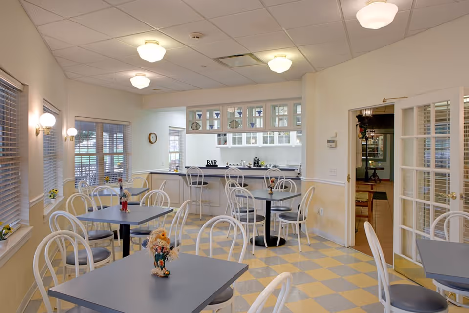 A bright dining area with multiple square tables and white chairs with gray cushions. The room has large windows with blinds, wall-mounted light fixtures, and a checkered yellow and gray floor. In the background, there is a bar counter with high stools and glass cabinets above it. A doorway with glass-paneled double doors leads to another room.