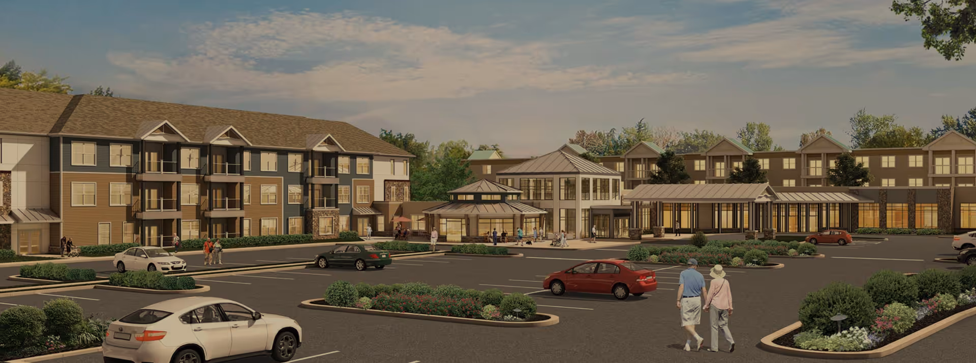 View of a senior living facility parking lot with several cars parked and a few people walking. The facility buildings are three stories tall with balconies and large windows, surrounded by landscaped greenery under a partly cloudy sky.