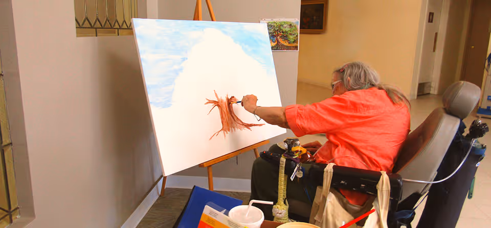 An elderly person in a wheelchair is painting a tree on a large canvas set on an easel in an indoor hallway or common area. The person is wearing a bright orange shirt and is focused on their artwork. Various personal items and art supplies are visible around the wheelchair.