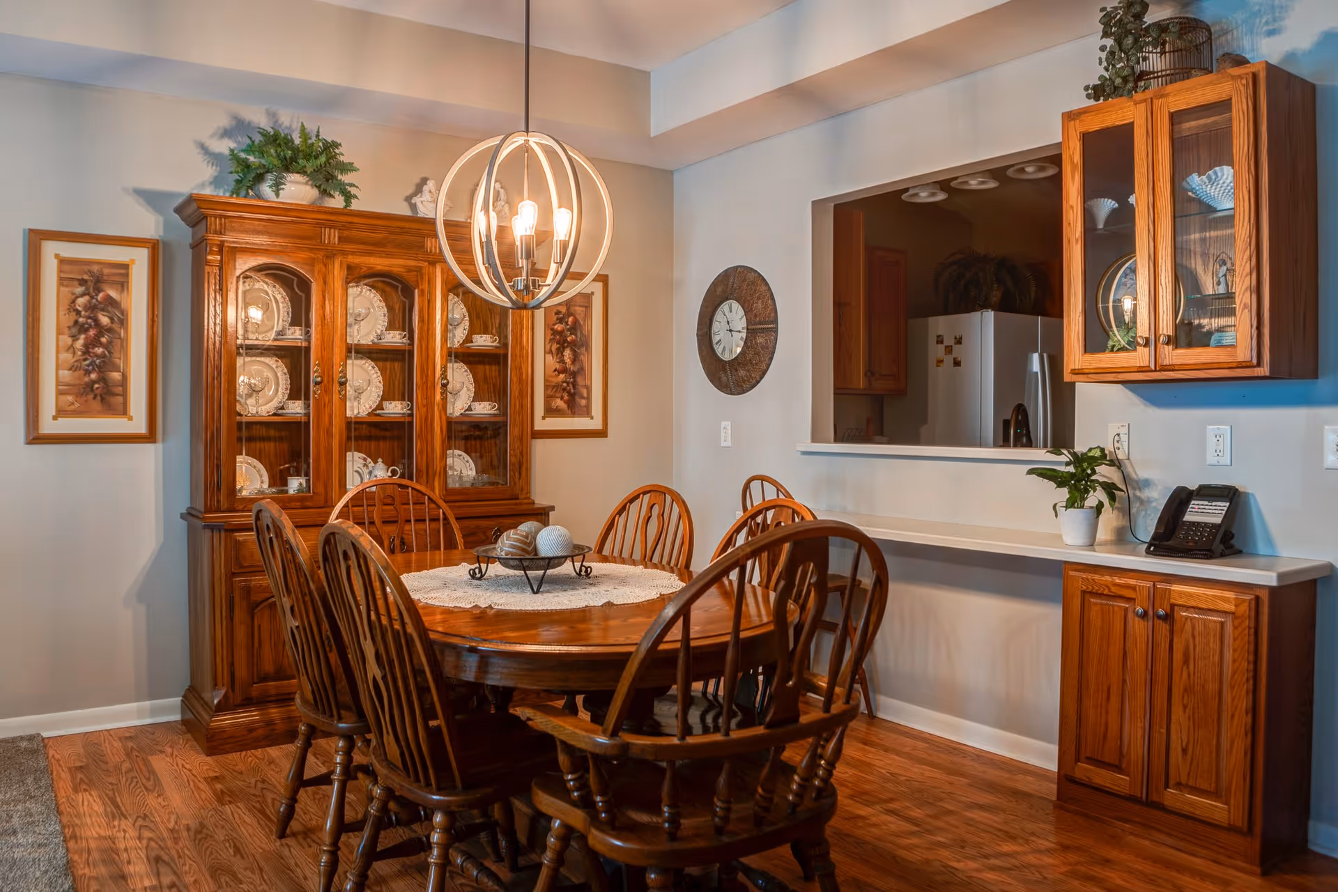 A dining room with a wooden dining table surrounded by six wooden chairs. A decorative centerpiece sits on a lace doily on the table. Behind the table is a wooden china cabinet displaying plates and teacups. The walls are light-colored with two framed pictures on either side of the cabinet. A modern spherical chandelier hangs above the table. To the right, there is a pass-through window to the kitchen, with a countertop below it and wooden cabinets on the wall and below the counter. A clock hangs on the wall near the pass-through, and a small potted plant and a phone are on the counter.
