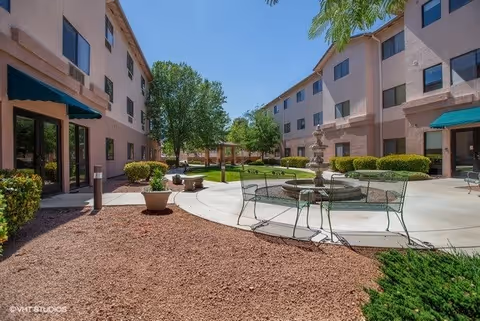 Outdoor courtyard area of a senior living facility with a central fountain surrounded by metal benches and chairs. The courtyard is bordered by two three-story buildings with windows and green awnings. There are trees, shrubs, and a clear blue sky overhead.