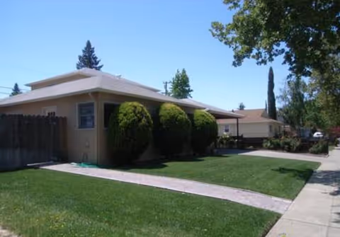 Single-story residential building with a beige exterior, surrounded by neatly trimmed bushes and a well-maintained green lawn. A concrete walkway leads to the entrance, and there are trees and other houses visible in the background under a clear blue sky.