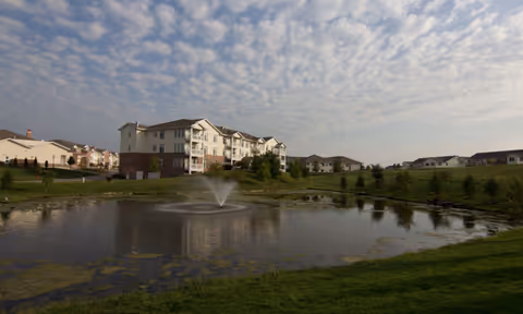 A large pond with a water fountain in the center, surrounded by green grass and trees. In the background, there are multiple multi-story residential buildings under a partly cloudy sky.