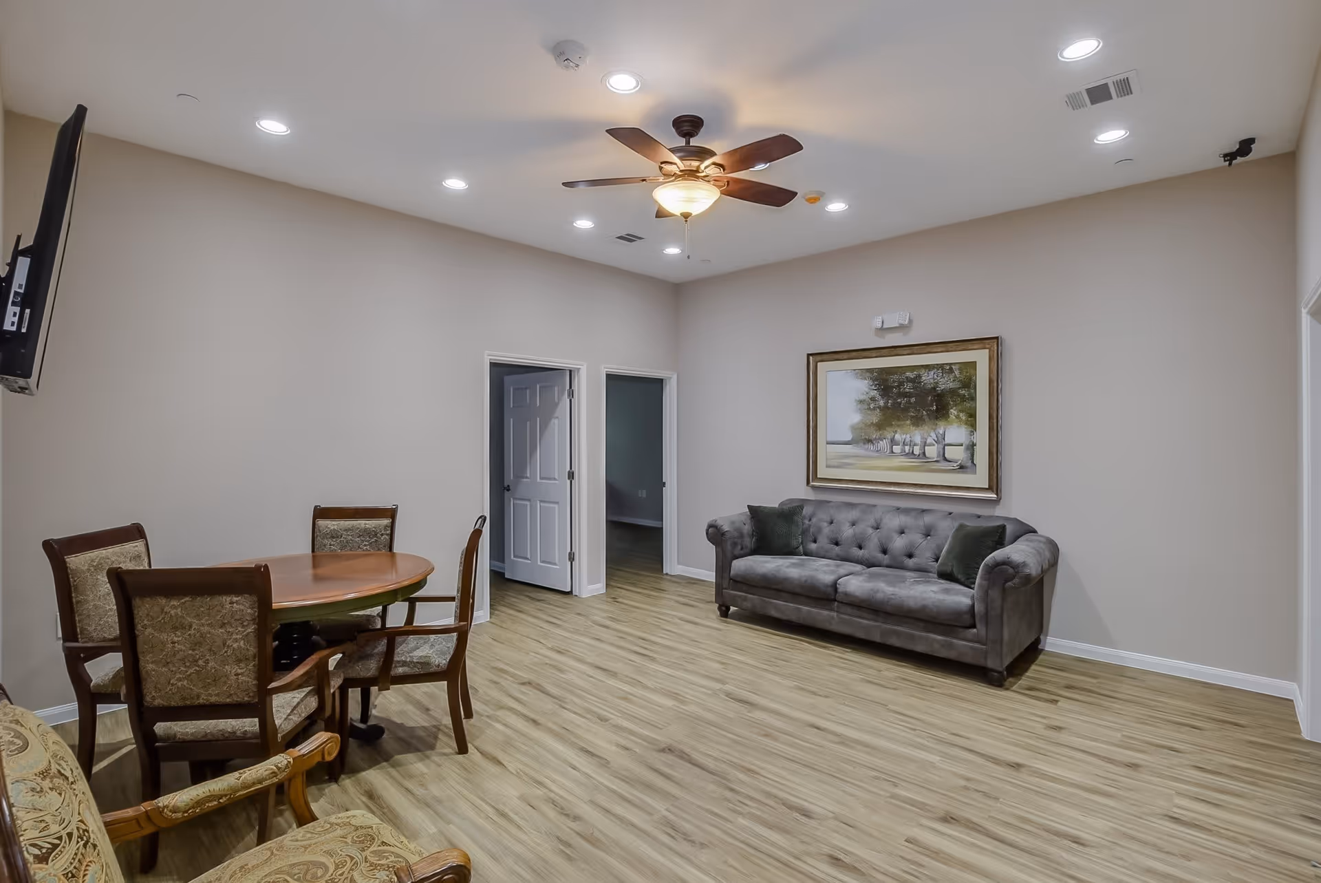 Community living area with a tufted gray sofa, round wooden table and chairs, ceiling fan, and framed artwork on the wall.