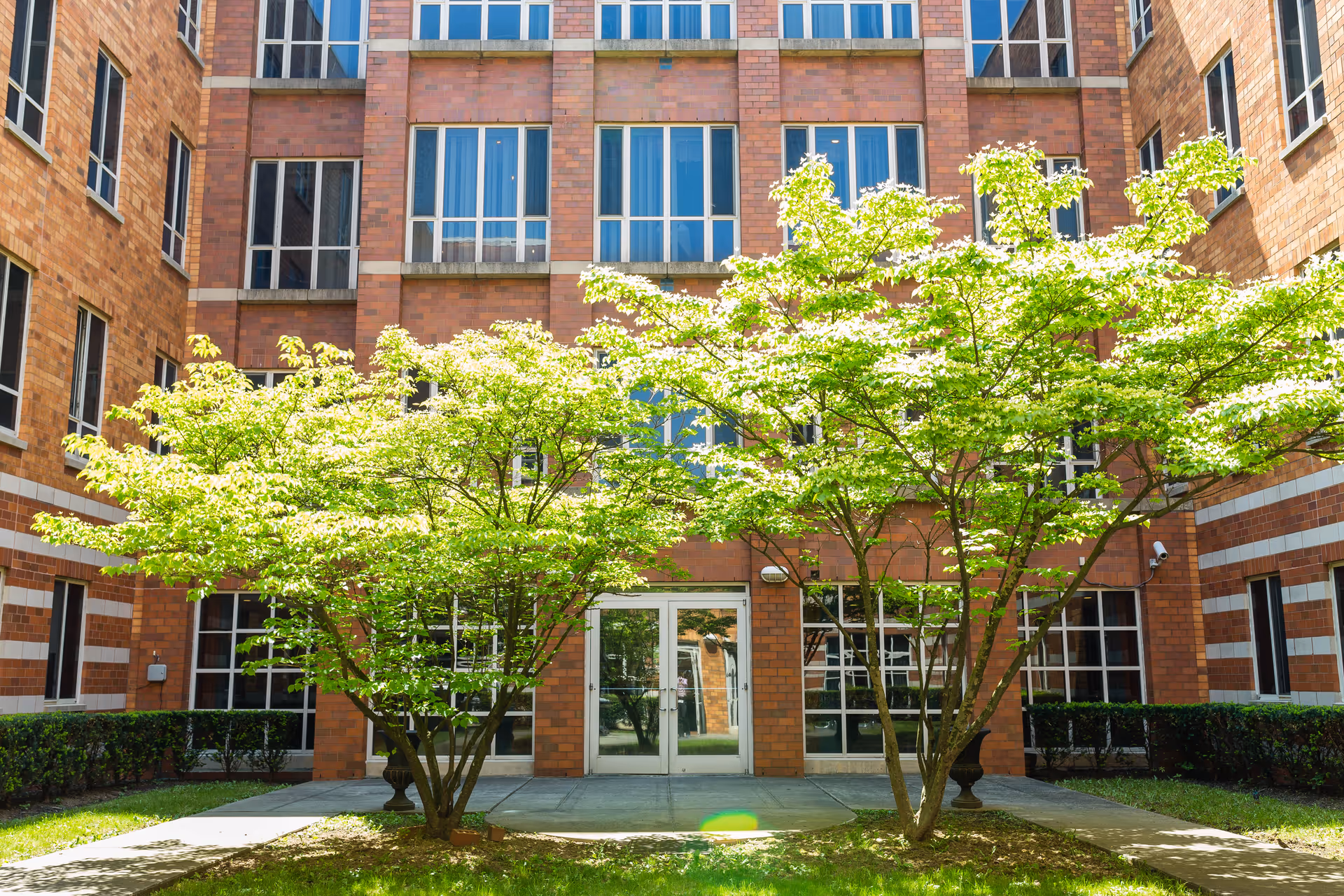Exterior view of a brick building with multiple windows and a glass door entrance. Two leafy green trees are in front of the entrance, with a small concrete walkway leading to the door. The building appears to be part of a senior living or nursing facility.