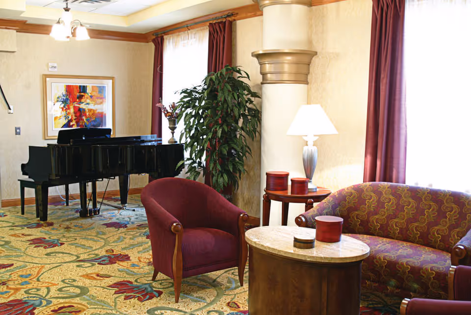 A cozy living room area with a patterned carpet, a maroon armchair, a matching maroon patterned loveseat, a round wooden coffee table with decorative items, a side table with a lamp, a large green plant, a grand piano, and a colorful abstract painting on the wall.