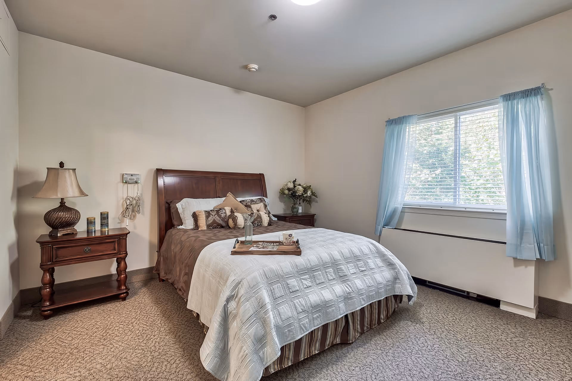 A neatly made bed with a wooden headboard in a bedroom. The bed has multiple pillows and a tray with a glass and a cup on it. There is a wooden nightstand with a lamp and decorative items on the left side of the bed. A window with light blue curtains lets in natural light on the right side of the room. The walls are painted light beige and the floor is carpeted.