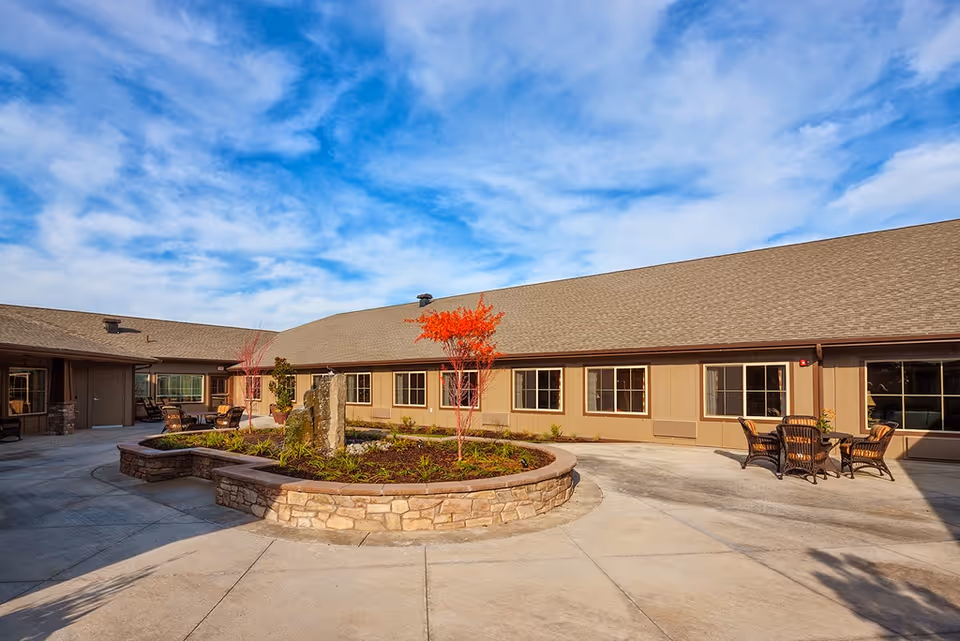 Outdoor courtyard area of The Hampton at Salmon Creek Memory Care Community featuring a circular stone planter with small trees and plants, surrounded by a paved walkway. The building has beige siding with multiple windows and a brown shingled roof. There are several outdoor seating areas with tables and chairs. The sky is partly cloudy with blue patches.