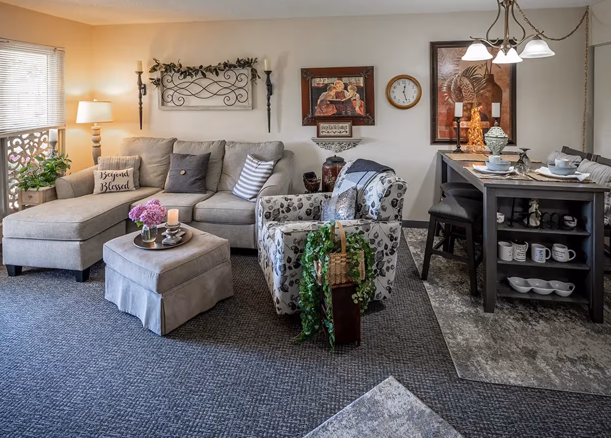 A cozy living room area with a beige sectional sofa adorned with multiple pillows, including one that says 'Beyond Blessed.' There is a matching ottoman with a tray holding a candle and flowers. Next to the sofa is a patterned armchair with a basket of greenery beside it. The wall behind the sofa features decorative wall sconces, framed artwork, and a clock. To the right, a dining table set for four with chairs and various decorative items is visible under a hanging light fixture.