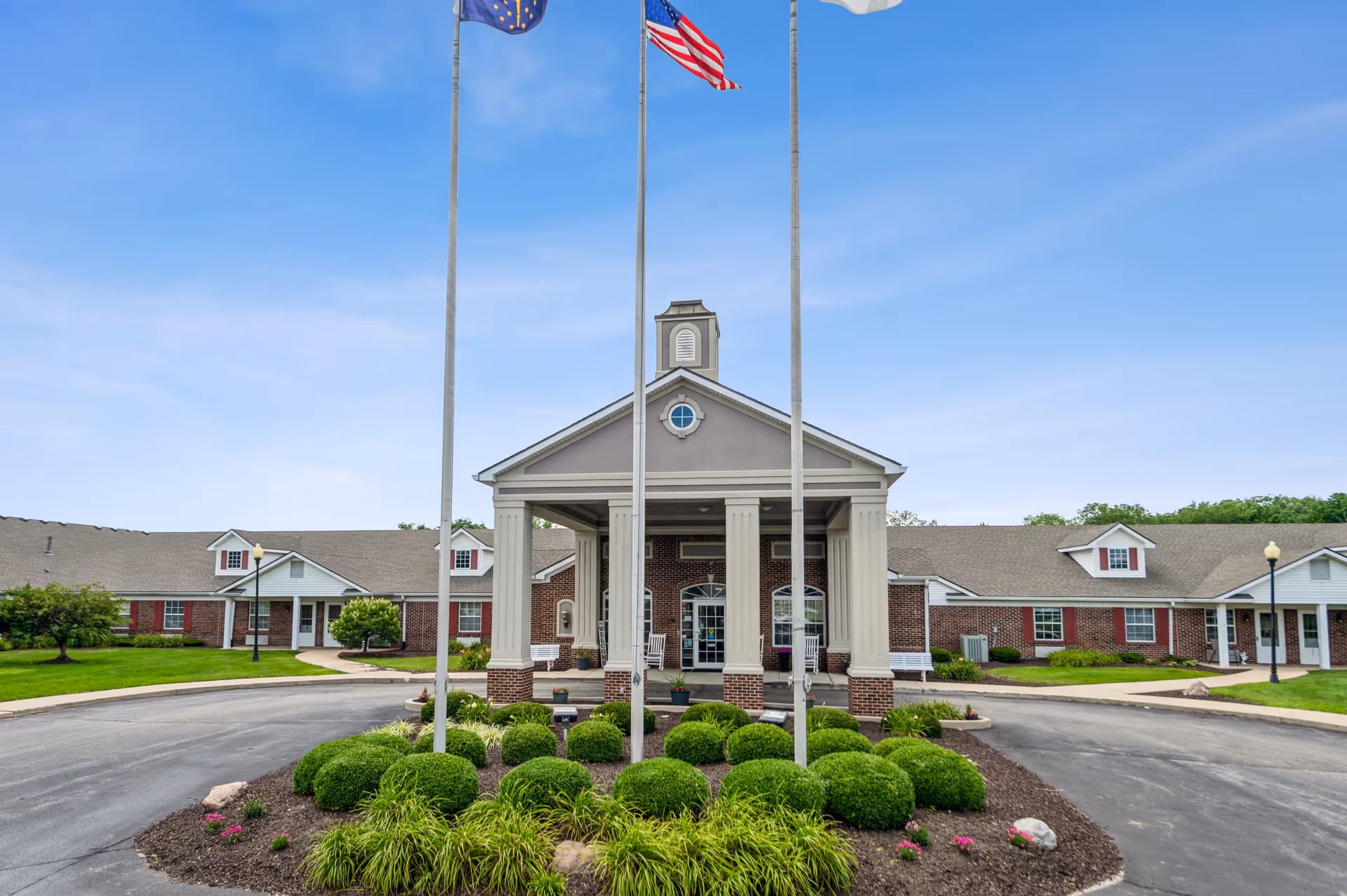 Front exterior of a senior living facility with a columned entrance portico, three flagpoles, circular driveway, and landscaped shrubs.