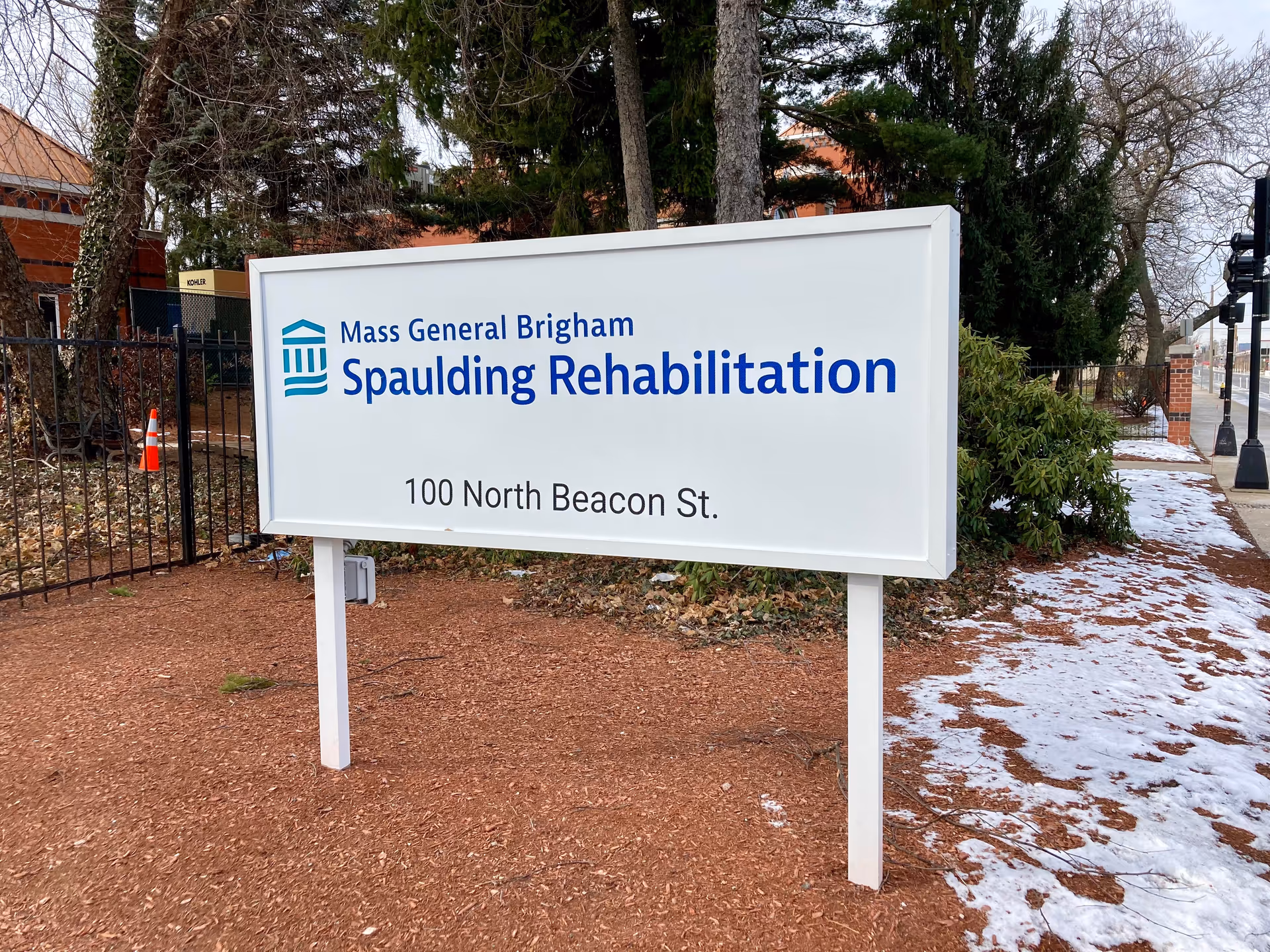 Outdoor sign for Mass General Brigham Spaulding Rehabilitation located at 100 North Beacon St., surrounded by trees, a fence, and patches of snow on the ground.