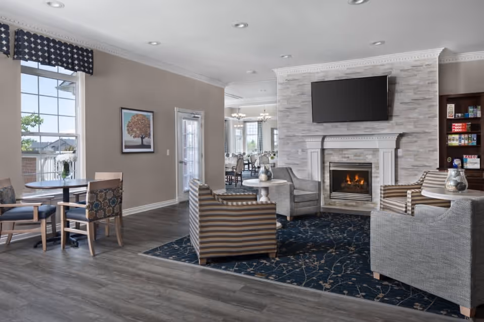 A cozy living room area in Waltonwood Lakeside featuring a fireplace with a mounted TV above it, surrounded by comfortable armchairs and small tables. To the left, there is a small dining area with a round table and chairs near a window with a patterned valance. The room has wood flooring and neutral-colored walls, with a glimpse of a larger dining area in the background.